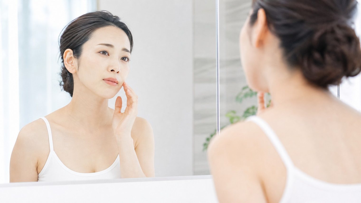 洗面所の鏡の前で頬の肌の調子をそっと確かめる30代前後の女性のイメージ写真。