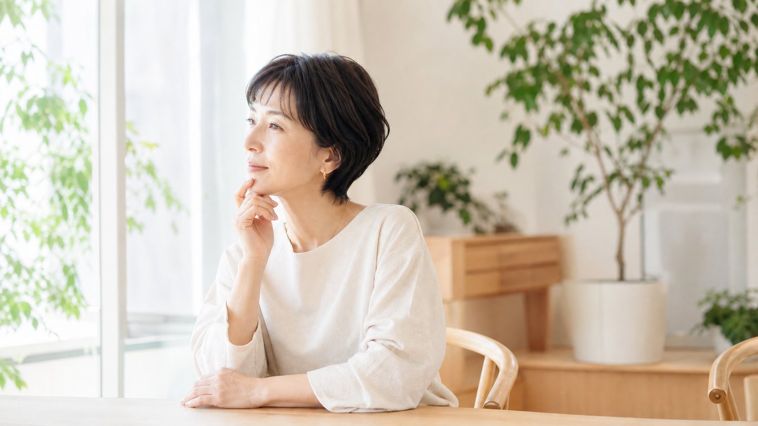 窓辺で少し疲れた表情をしている50代前後の女性。体調の変化が気になる場面のイメージ写真。
